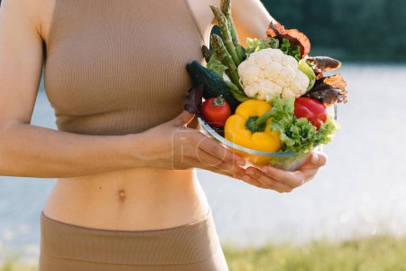 Photo for Female vegan holding plate of fresh vegetables near the belly in outdoors - Royalty Free Image