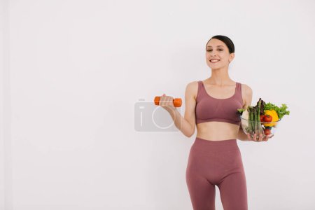 Téléchargez les photos : Belle jeune femme heureuse avec plateau de divers légumes sains et montrant ses muscles avec haltère isolé sur le fond blanc - en image libre de droit