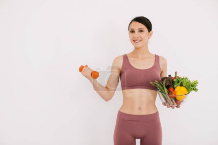 Photo for Beautiful happy young woman with tray of various healthy vegetables and showing her muscles with dumbbell isolated on the white background - Royalty Free Image