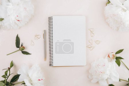 Foto de Espacio de trabajo femenino con bloc de notas abierto en blanco y flores de peonía, elegantes artículos de escritura de oficina y en un teble pastel - Imagen libre de derechos