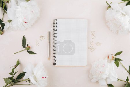 Foto de Espacio de trabajo femenino con bloc de notas abierto en blanco y flores de peonía, elegantes artículos de escritura de oficina y en un teble pastel - Imagen libre de derechos