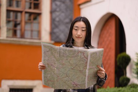 Téléchargez les photos : Portrait d'une belle brune coréenne heureuse tenant une carte dans les rues de la vieille ville. Femme asiatique touristique dame voyageant en Europe. - en image libre de droit