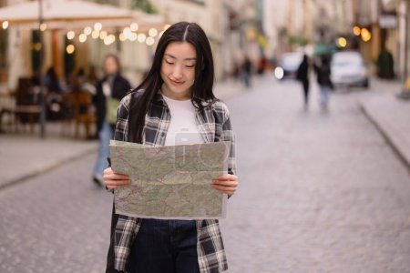 Téléchargez les photos : Portrait d'une belle brune coréenne tenant une carte dans les rues de la vieille ville. Femme asiatique touriste ou d'affaires dame voyageant en Europe. - en image libre de droit