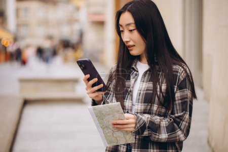 Téléchargez les photos : Portrait d'une belle brune coréenne tenant une carte et la comparant à la navigation par smartphone dans les rues de la vieille ville. Asiatique touristique femme voyageant en Europe. - en image libre de droit