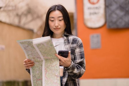 Photo for Portrait of a beautiful brunette Korean woman holding a map and comparing it with smartphone navigation on the streets of the old city. Asian tourist woman traveling in Europe. - Royalty Free Image