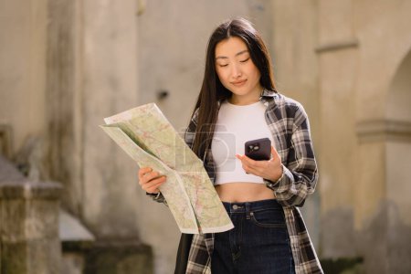 Téléchargez les photos : Portrait d'une belle brune coréenne tenant une carte et la comparant à la navigation par smartphone dans les rues de la vieille ville. Asiatique touristique femme voyageant en Europe. - en image libre de droit