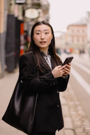 Photo for Young busy successful beautiful Asian business woman, korean professional businesswoman holding cellphone using smartphone standing or walking on old city street outside. - Royalty Free Image