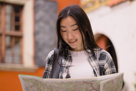 Téléchargez les photos : Portrait d'une belle brune coréenne heureuse tenant une carte dans les rues de la vieille ville. Femme asiatique touristique dame voyageant en Europe. - en image libre de droit