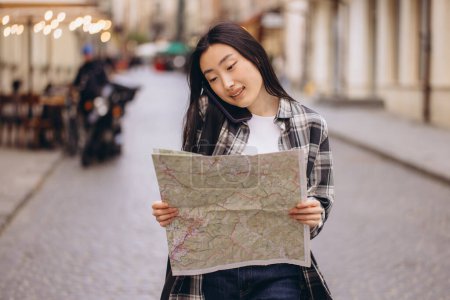 Foto de Retrato de una hermosa morena coreana sosteniendo un mapa y hablando por teléfono en las calles de la ciudad vieja. Turista asiática viajando por Europa. - Imagen libre de derechos