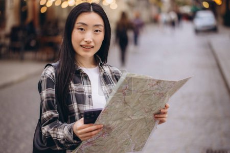 Photo for Portrait of a beautiful brunette Korean woman holding a map on the streets of the old city. Asian woman tourist or business lady traveling in Europe. - Royalty Free Image