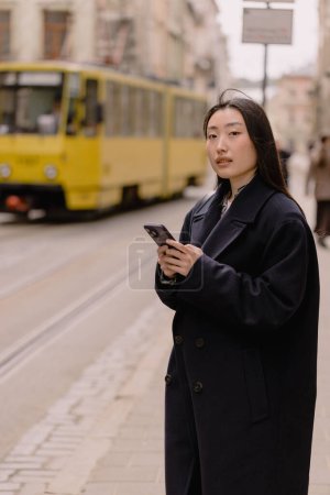 Foto de Retrato emocional de una mujer coreana en un abrigo sosteniendo un teléfono en una parada de transporte de la ciudad en el fondo de un tranvía y la arquitectura. - Imagen libre de derechos