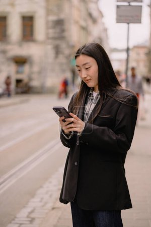 Photo for Young busy successful beautiful Asian business woman, korean professional businesswoman holding cellphone using smartphone standing or walking on old city street outside. - Royalty Free Image