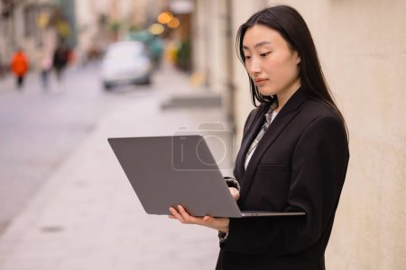 Foto de Retrato de una hermosa morena coreana con un portátil. Asiática mujer de negocios en un traje utiliza tecnología moderna. - Imagen libre de derechos