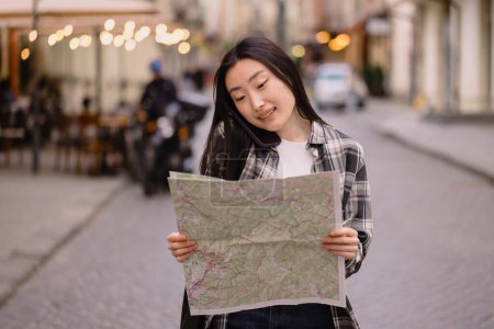 Téléchargez les photos : Portrait d'une belle brune coréenne tenant une carte et parlant au téléphone dans les rues de la vieille ville. Asiatique touriste femme voyageant en Europe. - en image libre de droit