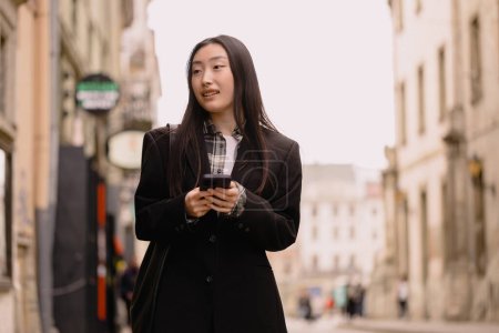 Téléchargez les photos : Jeune femme d'affaires asiatique couronnée de succès occupé belle, coréen femme d'affaires professionnelle tenant téléphone portable en utilisant smartphone debout ou marcher sur la rue de la vieille ville à l'extérieur. - en image libre de droit