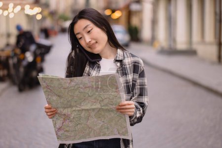 Foto de Retrato de una hermosa morena coreana sosteniendo un mapa y hablando por teléfono en las calles de la ciudad vieja. Turista asiática viajando por Europa. - Imagen libre de derechos