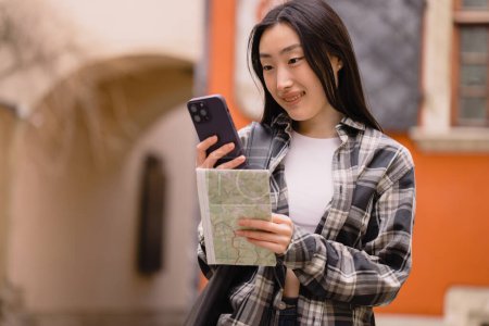 Photo for Portrait of a beautiful brunette Korean woman holding a map and comparing it with smartphone navigation on the streets of the old city. Asian tourist woman traveling in Europe. - Royalty Free Image