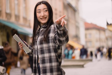 Téléchargez les photos : Portrait d'une belle brune coréenne tenant une carte et montrant quelque chose dans les rues de la vieille ville. Touriste asiatique voyageant en Europe. Espace de copie. - en image libre de droit