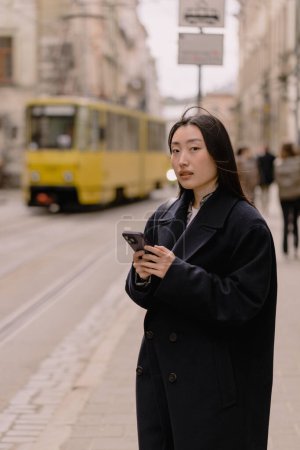 Foto de Retrato emocional de una mujer coreana en un abrigo sosteniendo un teléfono en una parada de transporte de la ciudad en el fondo de un tranvía y la arquitectura. - Imagen libre de derechos