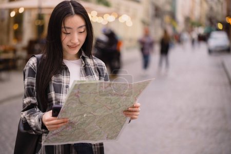 Photo for Portrait of a beautiful brunette Korean woman holding a map on the streets of the old city. Asian woman tourist or business lady traveling in Europe. - Royalty Free Image
