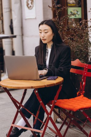 Téléchargez les photos : Entreprise indépendante jeune femme asiatique adulte utilisant un ordinateur portable pour le travail et parler par téléphone quelque chose. Assis à l'extérieur le jour. Style de vie des citadins avec la technologie moderne. - en image libre de droit