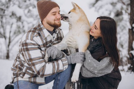 Foto de Feliz pareja joven con un perro akita en el bosque en invierno y día nevado. Un perro lame divertido oreja de maestro. - Imagen libre de derechos