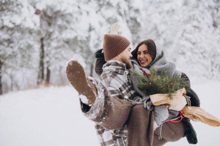 Photo for Cute young couple in love with pine bouquet spending time on Valentine's day in snowy winter forest in mountains. A guy holds a girl in his arms, smiling at each other. - Royalty Free Image