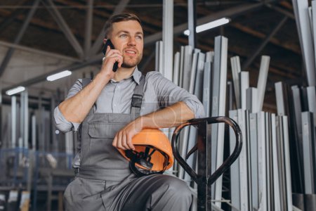 Photo for Portrait of a happy worker in a orange helmet and overalls holding a hydraulic truck and talking on the phone against a background of a factory and aluminum frames. - Royalty Free Image