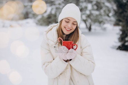 Téléchargez les photos : Mignonne femme heureuse en mitaines tricotées tenant du café chaud avec des cannes à bonbons dans le parc d'hiver - en image libre de droit