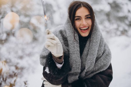 Foto de Retrato de mujer bonita feliz en bufanda de punto y manoplas que sostienen chispeante y posan en el bosque nevado - Imagen libre de derechos