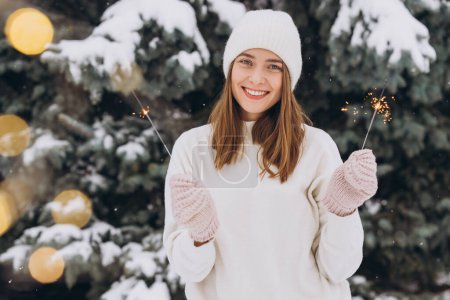Photo for Portrait of smiling girl wearing white knitted hat, scarf, mittens and sweater with sparkler in winter time outdoors. Christmas holidays - Royalty Free Image