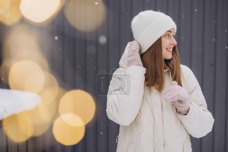 Téléchargez les photos : Une femme heureuse en mitaines tricotées pose sur un fond de mur gris en hiver dans une chute de neige - en image libre de droit