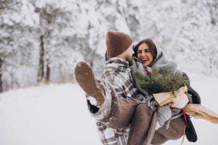 Photo for Cute young couple in love with pine bouquet spending time on Valentine's day in snowy winter forest in mountains. A guy holds a girl in his arms, smiling at each other. - Royalty Free Image