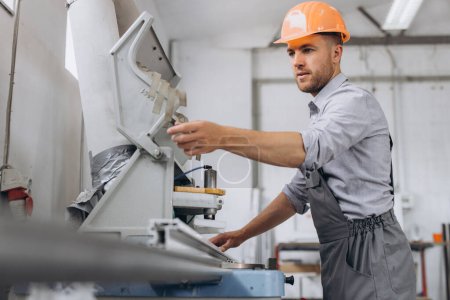 Téléchargez les photos : Un travailleur masculin en uniforme spécial et un casque orange travaillent sur une machine CNC pour la production de fenêtres et de portes en PVC à l'usine. - en image libre de droit