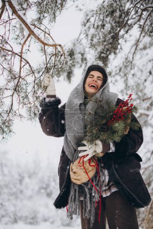 Foto de Retrato de mujer bonita feliz en bufanda de punto y mitones que sostienen ramo de pino de invierno y posan en el bosque nevado - Imagen libre de derechos