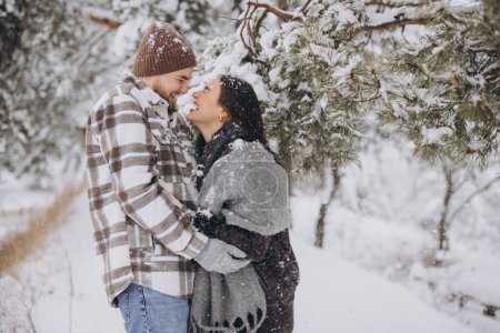 Photo for A young happy and loving couple is standing under a pine tree from which snow falls in the mountains in winter. - Royalty Free Image