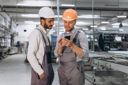 Photo for Two international male workers in overalls looking at the phone against the background of the production of aluminum PVC windows and doors - Royalty Free Image
