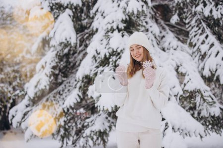 Photo for Happy woman in knitted white clothes holding snowflakes on the background of a snowy pine tree in winter - Royalty Free Image