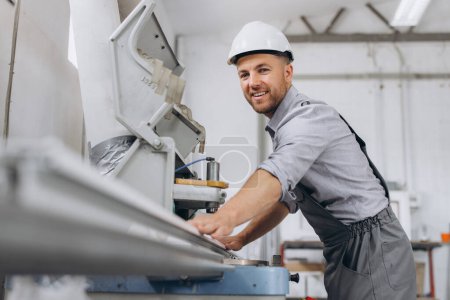 Téléchargez les photos : Un travailleur masculin en uniforme spécial et un casque blanc travaillent sur une machine CNC pour la production de fenêtres et de portes en PVC à l'usine. - en image libre de droit