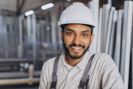 Photo for Portrait of a happy Hindu worker in a white hard hat and overalls holding a hydraulic truck against a background of a factory and aluminum frames. - Royalty Free Image