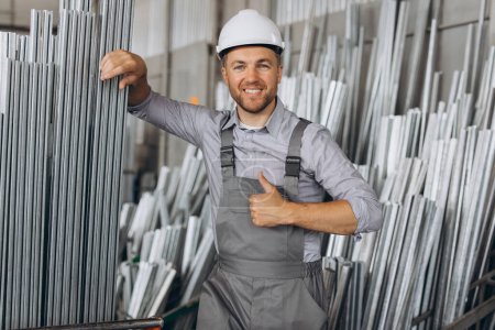 Photo for Happy bearded male factory worker in special uniform and white hard hat holding aluminum frame at production of metal plastic windows and doors - Royalty Free Image