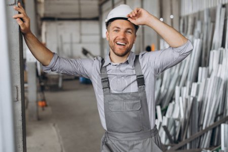 Photo for Portrait of a happy worker in a gray uniform and a white hard hat posing on the background of factory production - Royalty Free Image