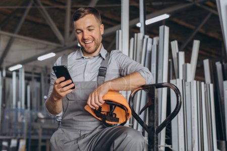 Foto de Retrato de un trabajador feliz en un casco naranja y overoles sosteniendo un camión hidráulico y hablando por teléfono sobre un fondo de una fábrica y marcos de aluminio. - Imagen libre de derechos