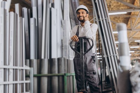 Photo for Portrait of a happy Hindu worker in a white hard hat and overalls holding a hydraulic truck against a background of a factory and aluminum frames. - Royalty Free Image