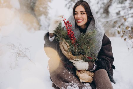 Foto de Retrato de mujer bonita feliz en bufanda de punto y mitones que sostienen ramo de pino de invierno y posan en el bosque nevado - Imagen libre de derechos