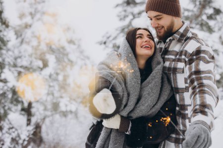 Téléchargez les photos : Couple romantique souriant en bonnets tricotés posant avec des étincelles à la forêt d'hiver, tenant les lumières du Bengale dans les mains, célébrant ensemble les vacances de Noël, Copier l'espace - en image libre de droit