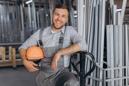Photo for Portrait of a happy worker in a orange hard hat and overalls holding a hydraulic truck against a background of a factory and aluminum frames. - Royalty Free Image