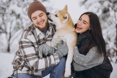 Foto de Feliz pareja joven con un perro akita en el bosque en invierno y día nevado. Un perro lame divertido oreja de maestro. - Imagen libre de derechos