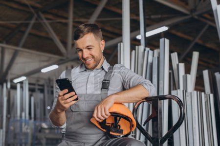 Foto de Retrato de un trabajador feliz en un casco naranja y overoles sosteniendo un camión hidráulico y hablando por teléfono sobre un fondo de una fábrica y marcos de aluminio. - Imagen libre de derechos