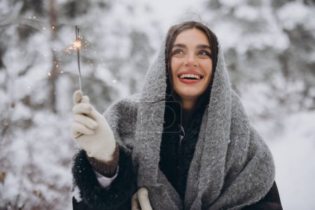 Photo for Portrait of happy pretty woman in knitted scarf and mittens holding sparkler and posing in snowy forest - Royalty Free Image